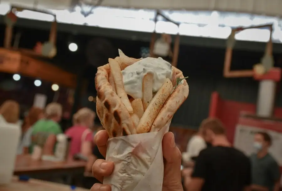 A hand holds a gyro wrapped in paper, topped with tzatziki sauce, in a food court setting.