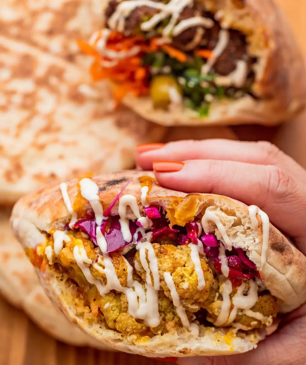 A hand holds a pita filled with chickpea patties, slaw, and tahini sauce, with another pita in the background.