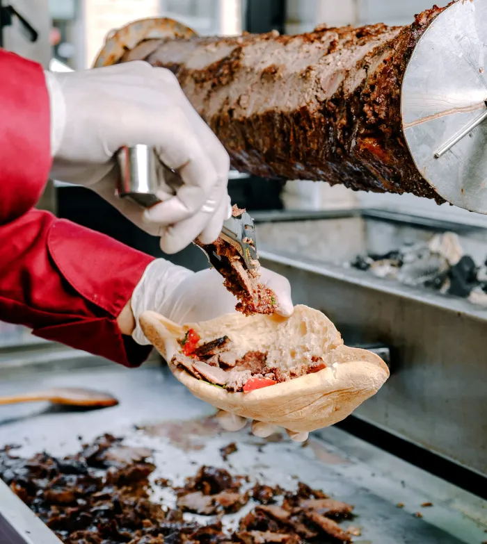 Cook shaving shawarma meat from a vertical spit onto pita bread with tomato and greens