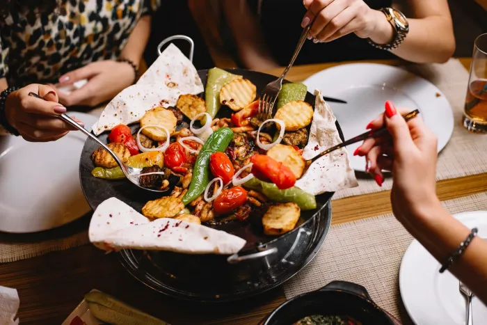 Diners sharing a skillet of grilled meat, potatoes, peppers, and tomatoes with flatbread