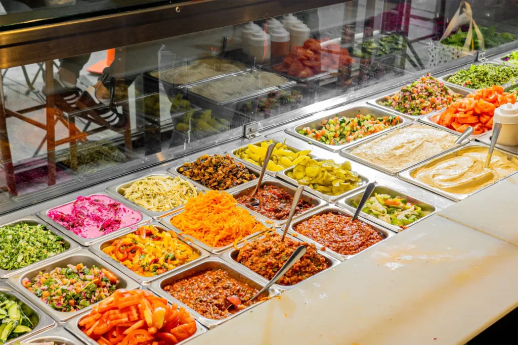 Salad and topping bar display with tabbouleh, carrots, tomatoes, hummus, pickles and Turkish spreads