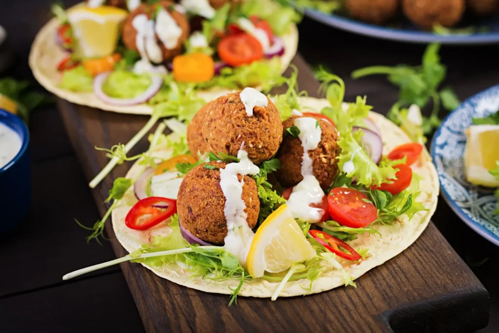 Falafel wraps with lettuce, tomatoes, onions, lemon, and tahini sauce on a wooden board.