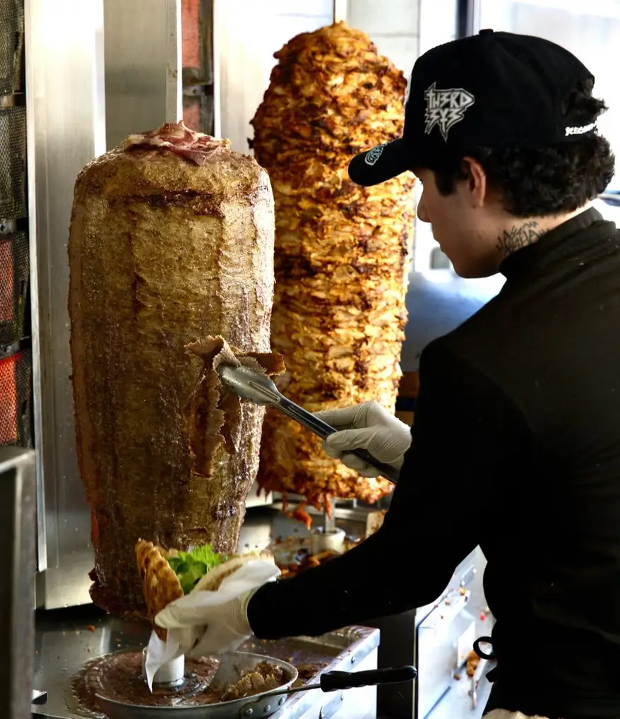 Staff member carving beef and chicken shawarma from the vertical rotisserie into fresh pita