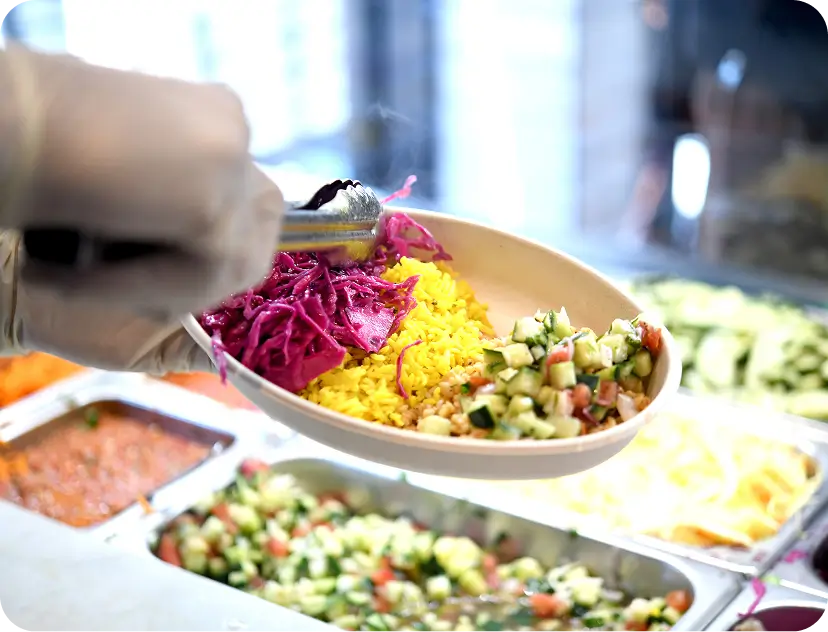 Gloved hand assembling a bowl with yellow rice, pickled red cabbage, and cucumber tomato salad at the counter