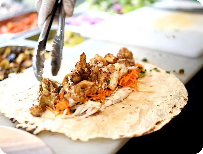 Gloved hand using tongs to add seasoned chicken shawarma onto a flatbread with shredded carrots and cabbage